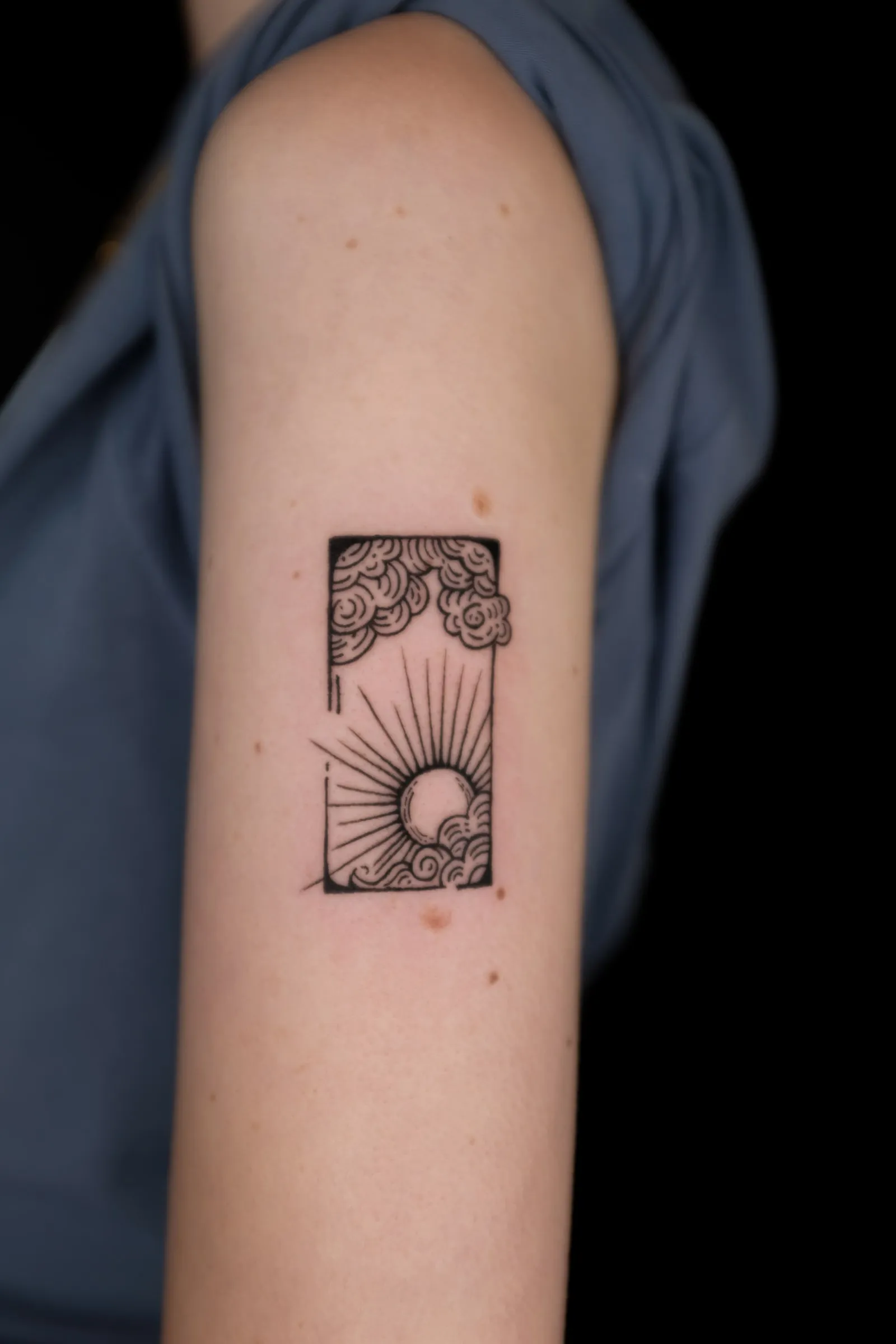 Finished upper-arm tattoo of a rising sun waves and layered clouds inside an open rectangular frame