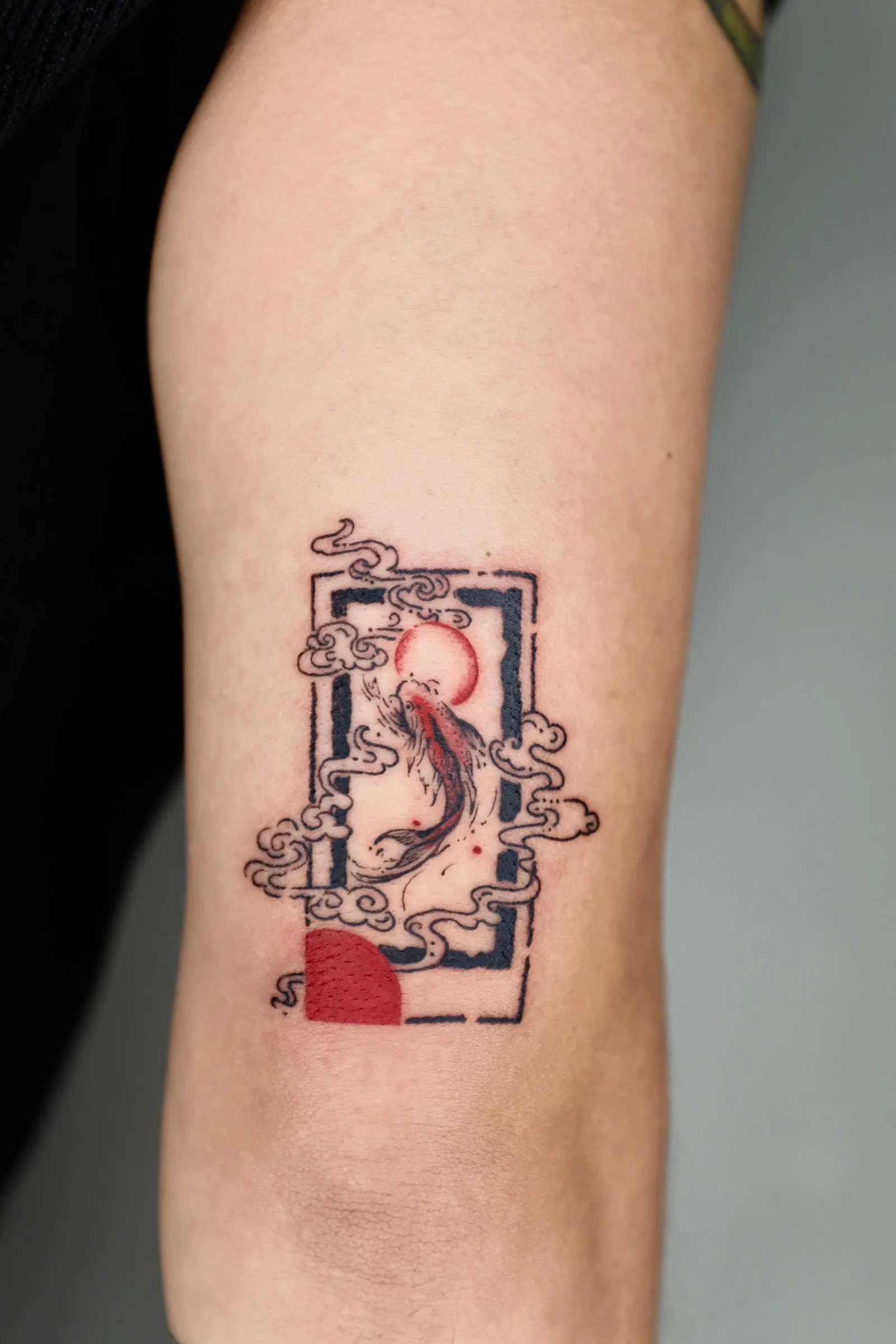 Finished arm tattoo of a red-and-black koi swimming through clouds inside a tall black frame