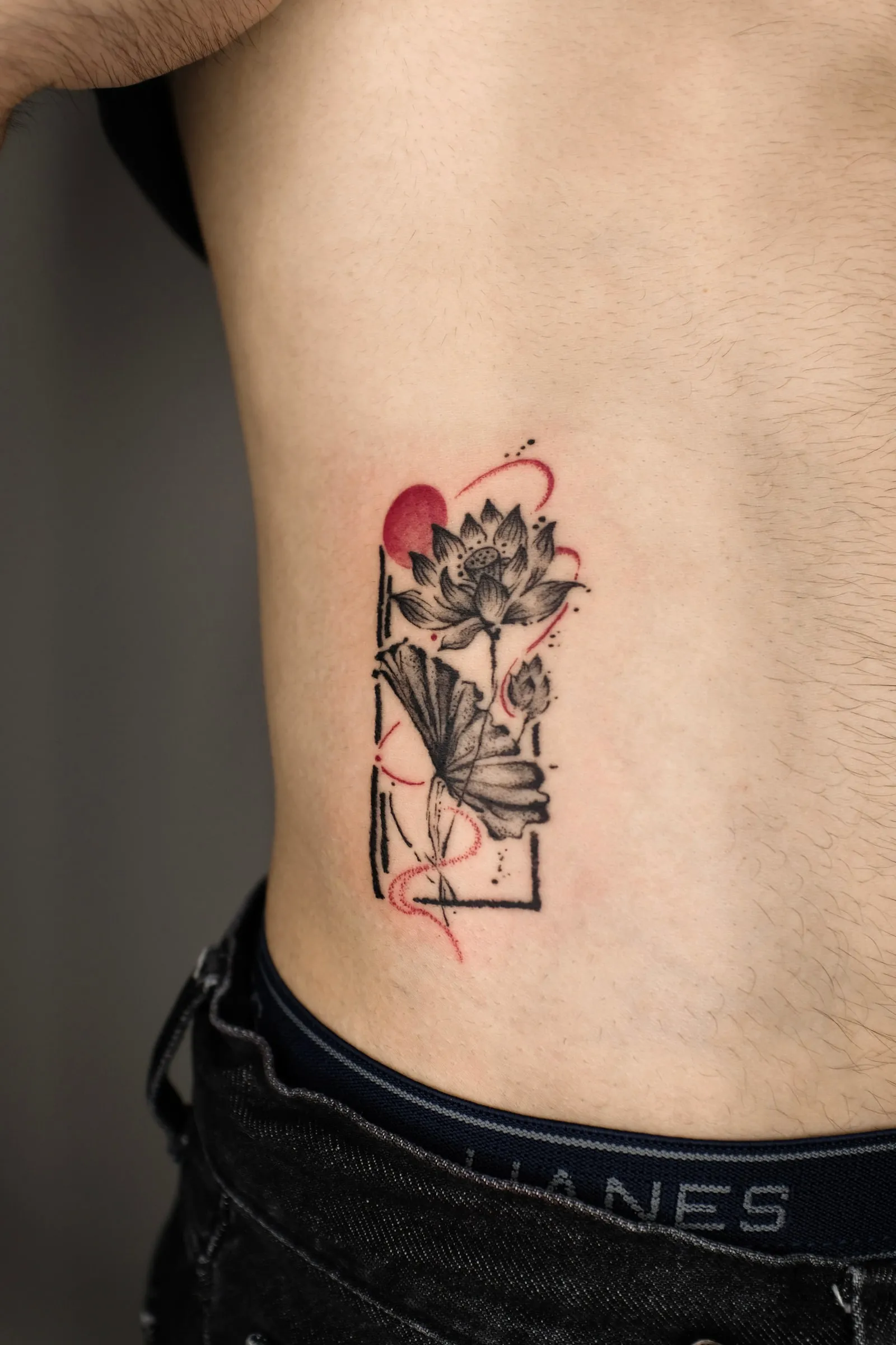 Finished rib tattoo of lotus and ginkgo leaves with red brush accents inside an open frame