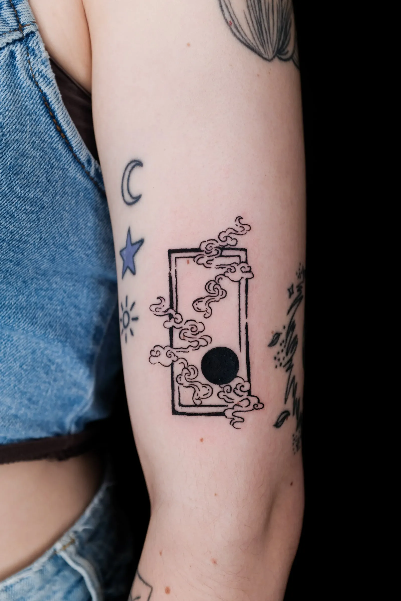 Finished arm tattoo of a black moon surrounded by curling clouds inside a tall open frame