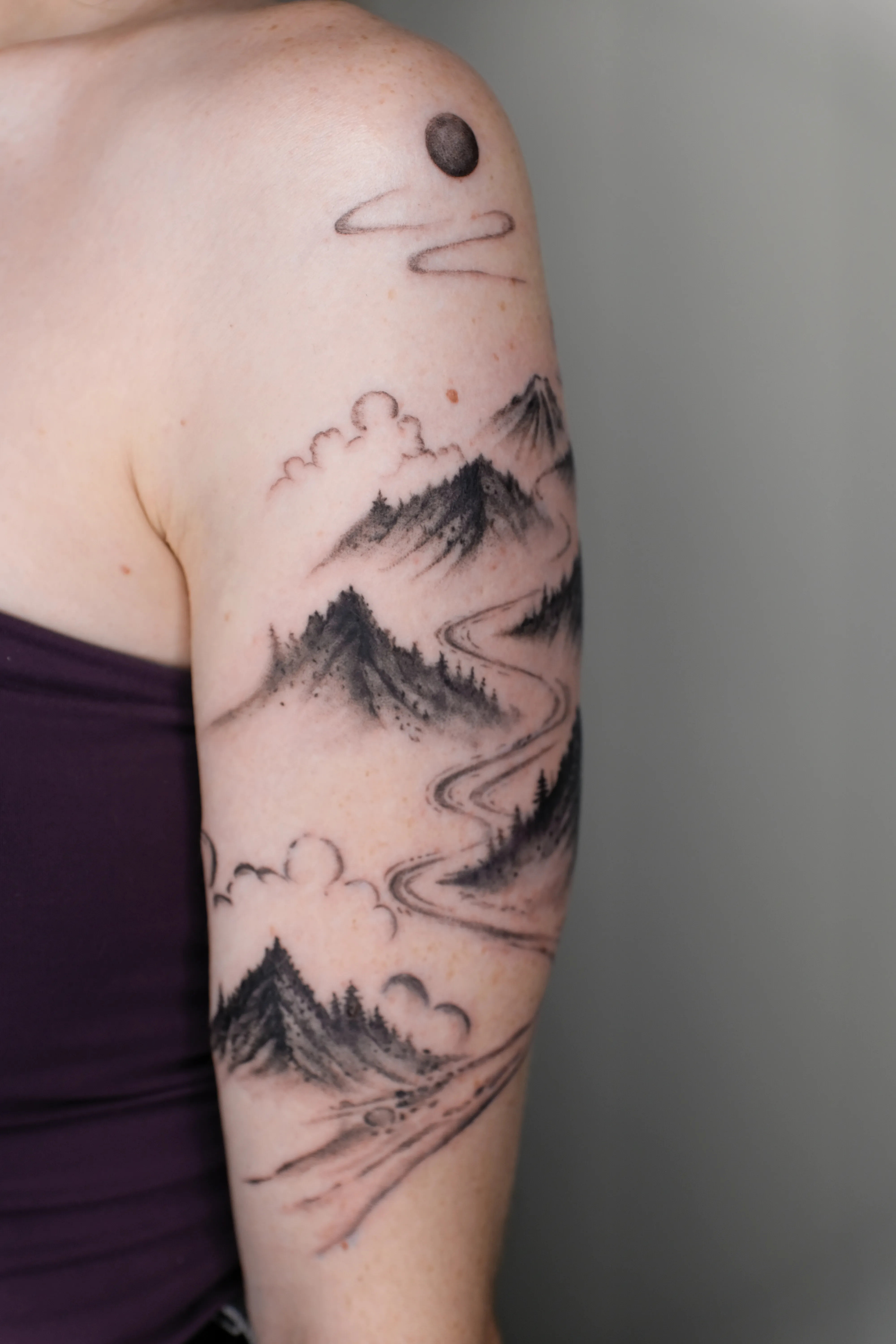 Finished upper-arm tattoo of dark mountain ridges clouds a black sun and a winding river