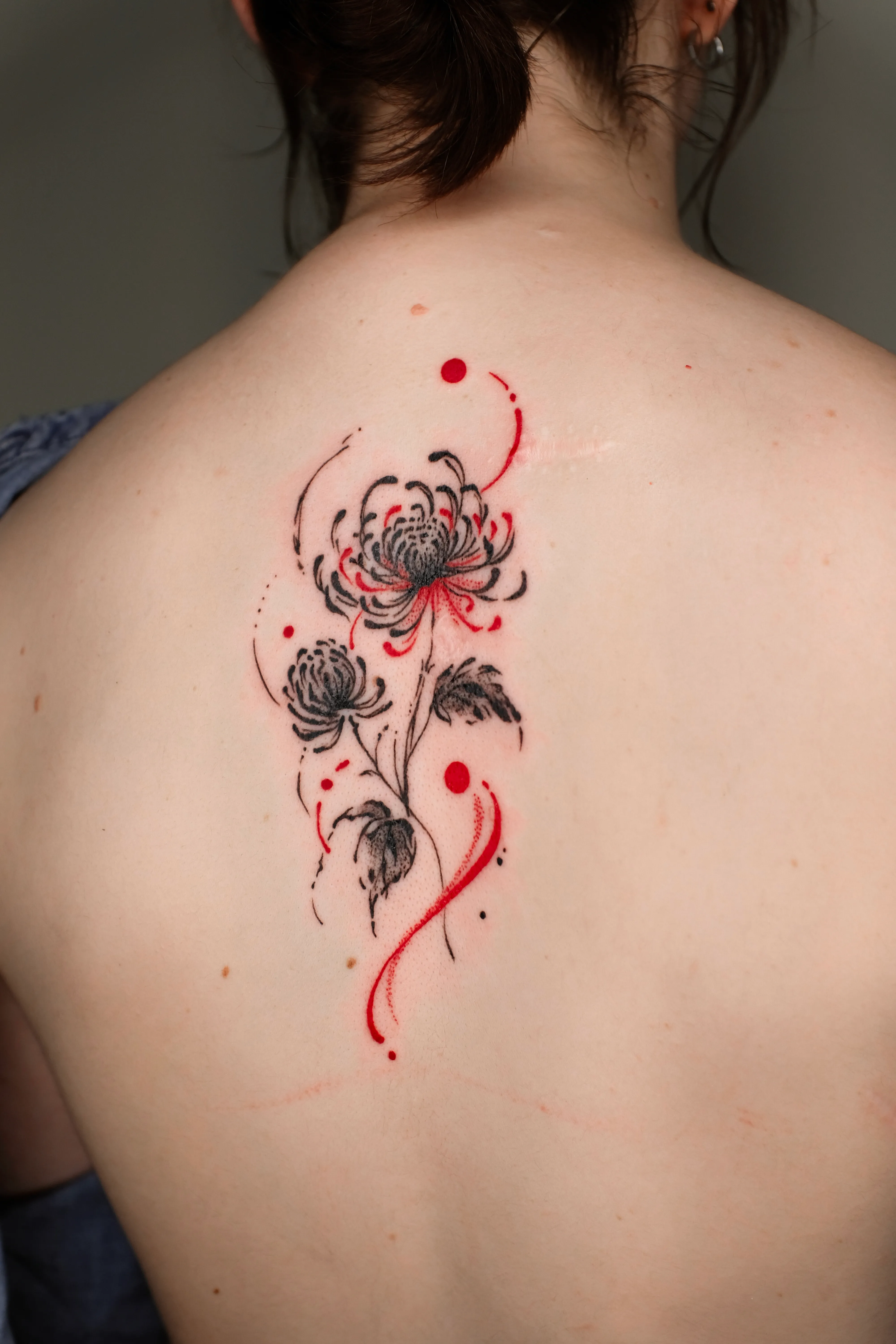 Finished upper-back tattoo of a black chrysanthemum with sweeping red brush marks and dots