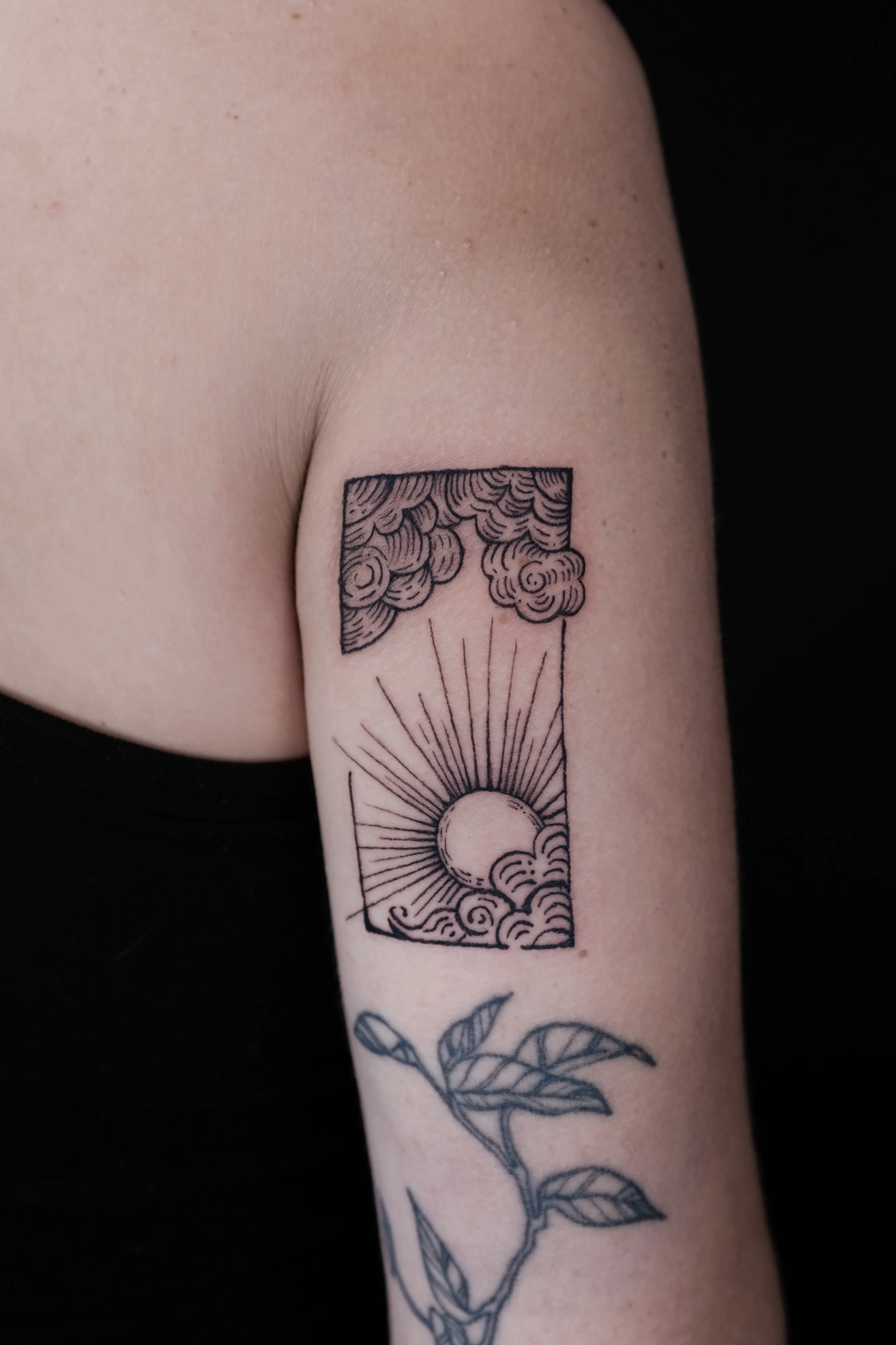 Finished upper-arm tattoo of a rising sun waves and layered clouds inside an open vertical frame
